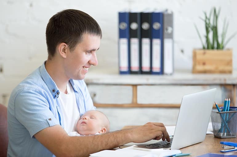 Side view portrait of happy casual young business dad holding newborn cute babe while working on laptop in home office interior. Cheerful working father using pc computer and nursing new born child