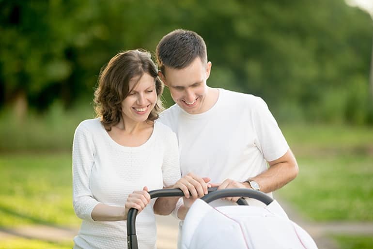 Happy young parents dressed in white clothes walking in park in summer with baby carriage. They standing and looking at baby sleeping in pram with love and care