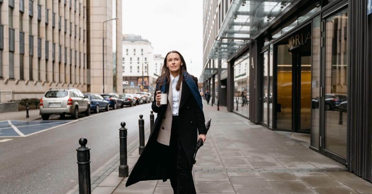 A woman walks along a city sidewalk holding a cup of coffee in soft morning light.