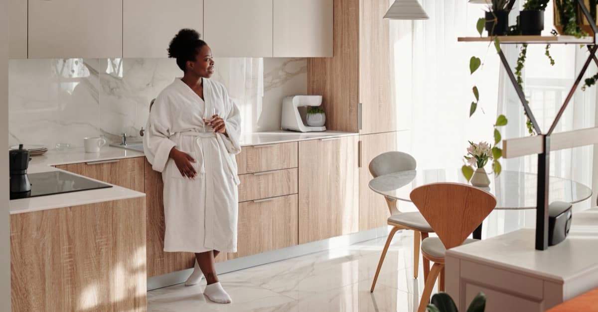 A woman in a white robe leans against a sunlit kitchen counter with a mug, calm in the early morning light.