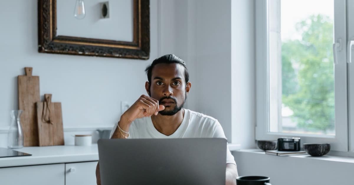 A man sits at a laptop in a bright, calm kitchen with a coffee cup nearby, hand resting on his chin in thought.