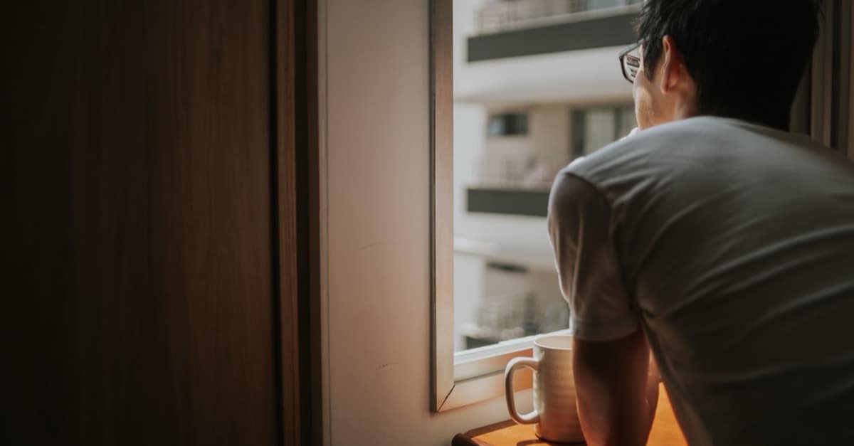 A man holds a coffee mug and looks thoughtfully out a window at city buildings in soft morning light.