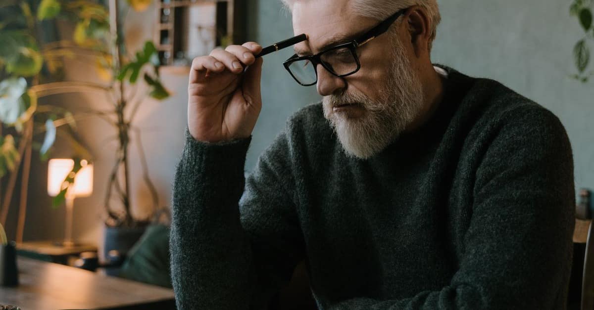 A thoughtful gray-haired man in his 50s pauses with his glasses raised, reflecting in warm, plant-filled home light.