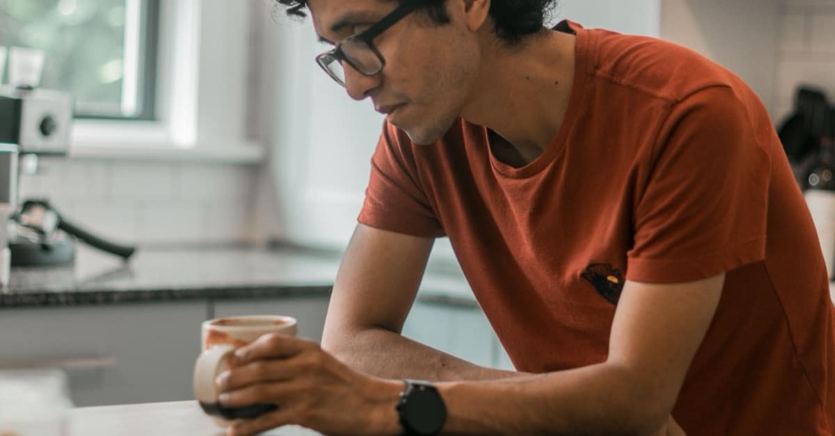 A man in glasses leans on a kitchen counter holding a coffee mug, looking down in calm thought in soft morning light.
