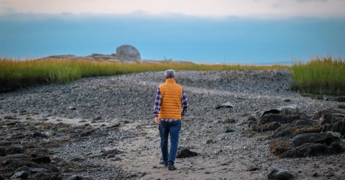 A grey-haired man in his fifties in an orange vest walks alone along an open gravel path toward the water at dawn, a sense of a new chapter and forward motion.