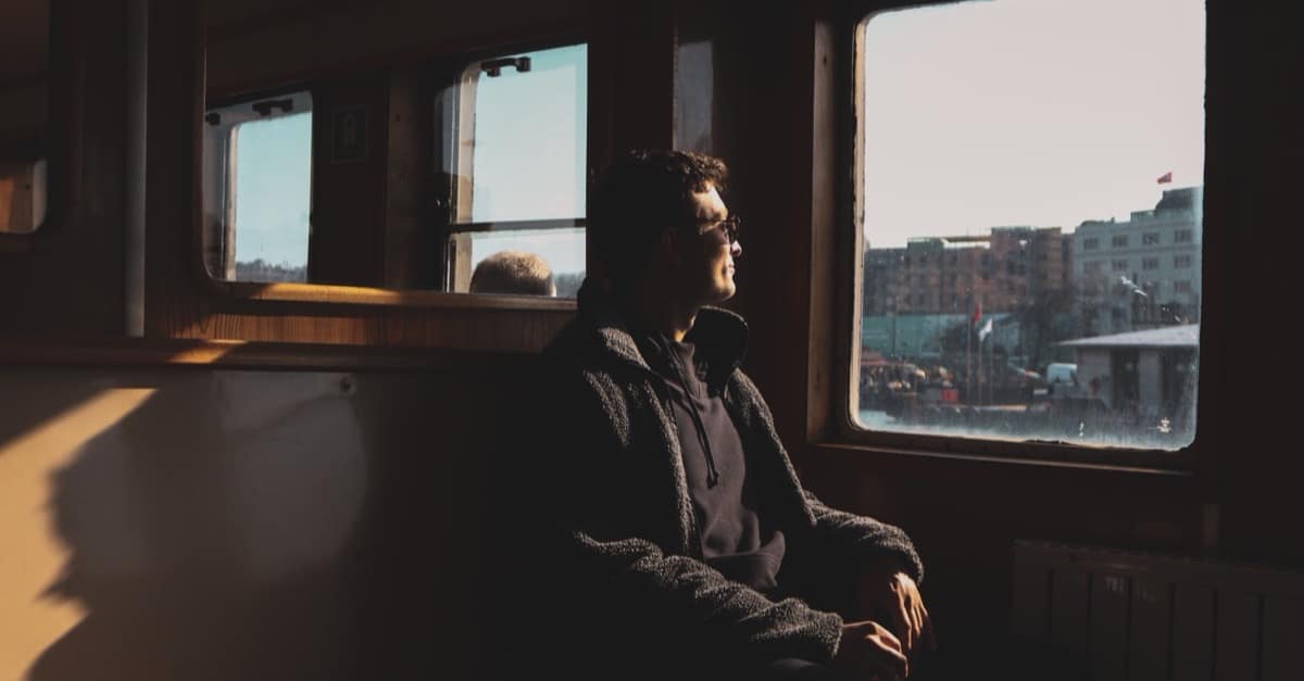 A man sits in contemplation, gazing out of a train window toward a city in soft light, with nothing in his hands.
