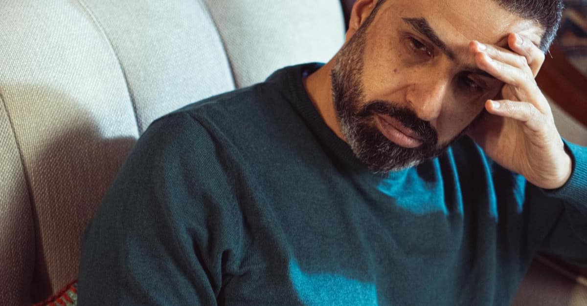 A man in his late 40s sits on a sofa with a hand to his forehead, tired but composed in warm indoor light.
