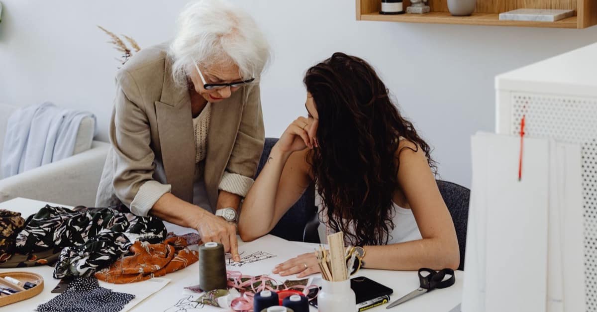 A woman with white hair leans over a bright studio worktable, pointing something out on a design sketch to a younger colleague sitting beside her.