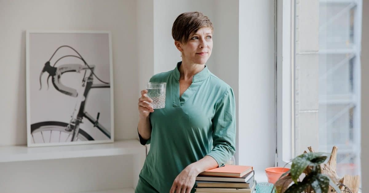 A woman in her late 40s stands at a bright window at home with a glass of water, looking out thoughtfully.