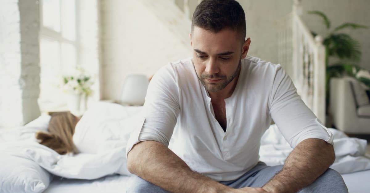A man in his late 40s sitting on the edge of an unmade bed in bright morning light, shirt half buttoned, looking tired but composed at the start of the day.