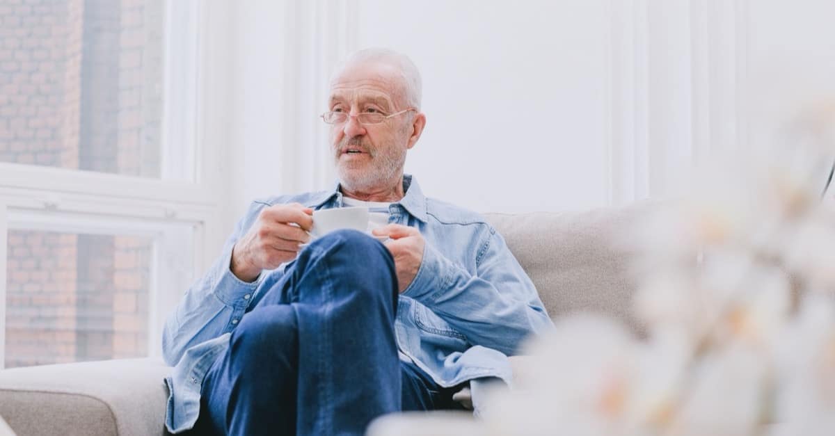 A man in his sixties sits with a cup of coffee in a bright, quiet living room on a weekday morning, looking away from the camera.