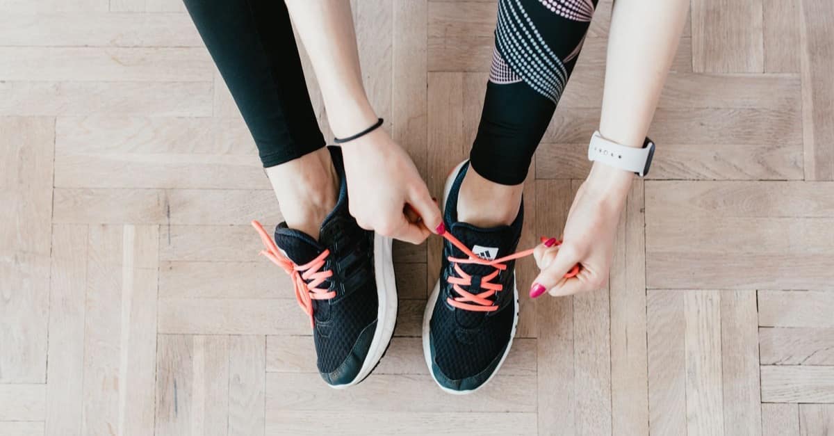 Overhead view of a woman in workout clothes lacing her sneakers on a light wooden floor at home in the morning.