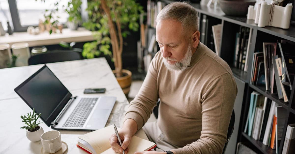 A man writing in an open notebook at a sunlit kitchen table, coffee and a closed laptop beside him.