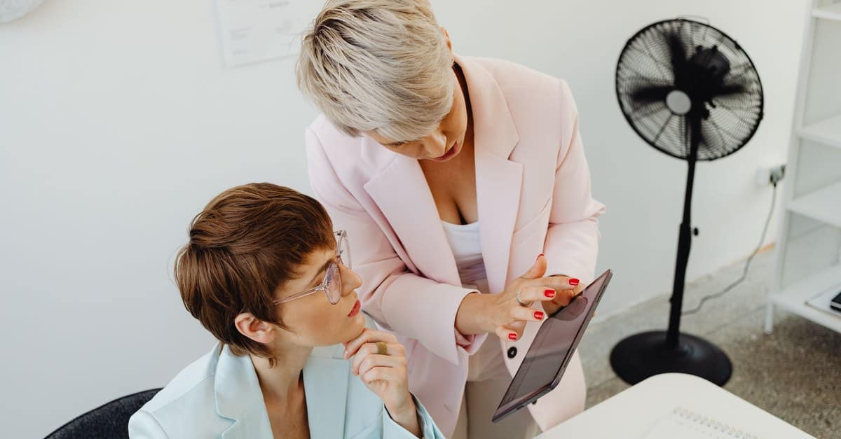 A colleague leans in with a tablet mid-request while a woman in her late forties looks up from her desk, already about to agree.