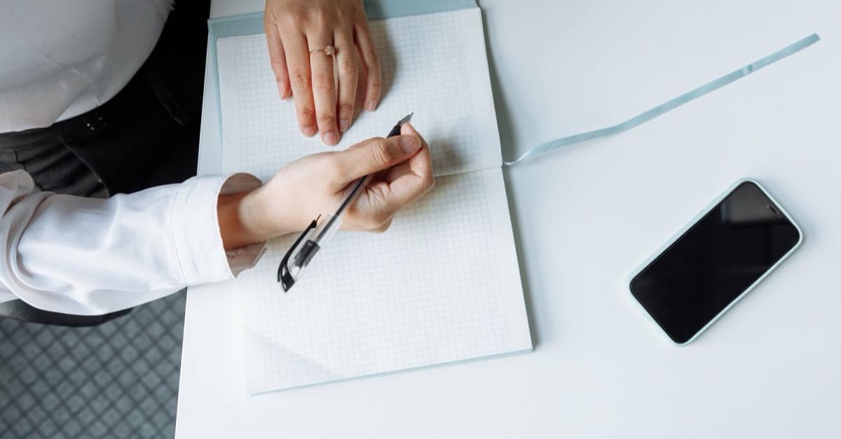 Overhead view of a person at a bright white table with an open grid notebook, one pen, and a phone set aside, ready to start a life audit.
