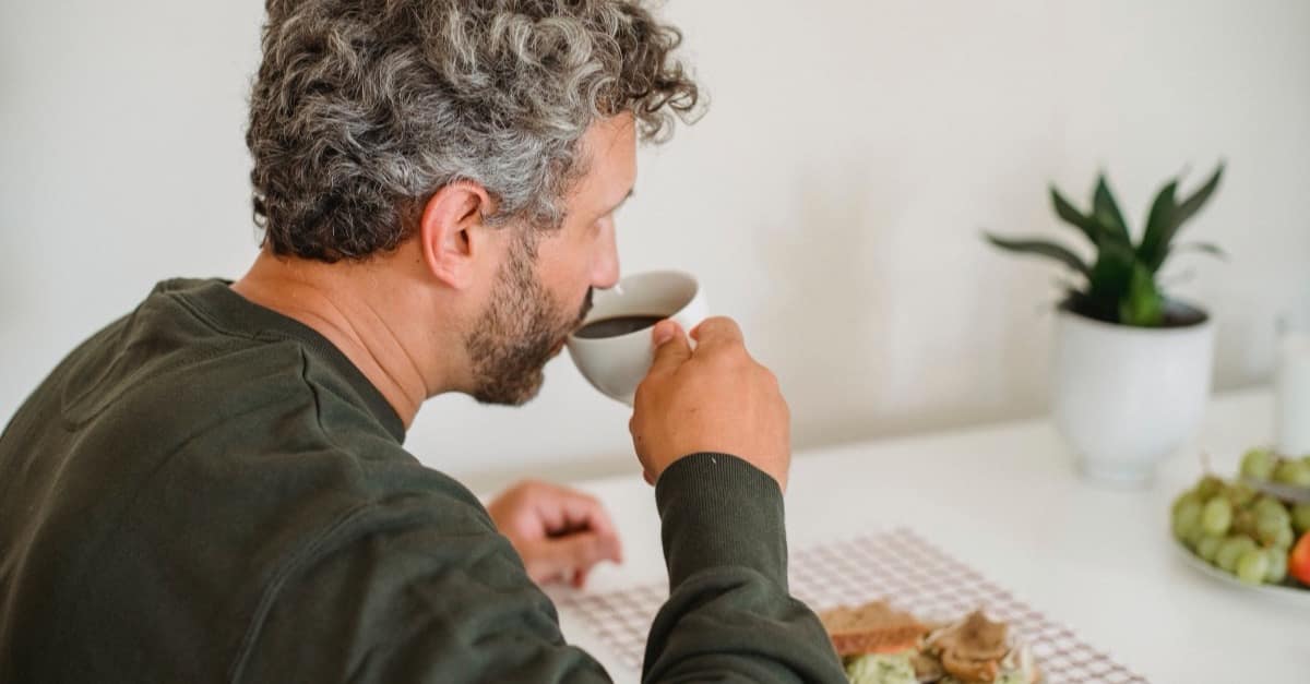 A man in his forties with graying hair pausing over a cup of coffee at a bright kitchen table, mid-morning, with nothing else on the table demanding him.