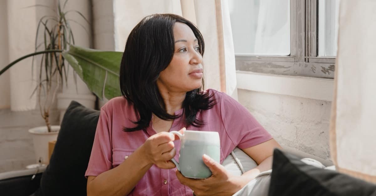 A woman in her forties sitting on a sofa by a bright window, holding a mug in both hands and looking out, with nothing scheduled in front of her.
