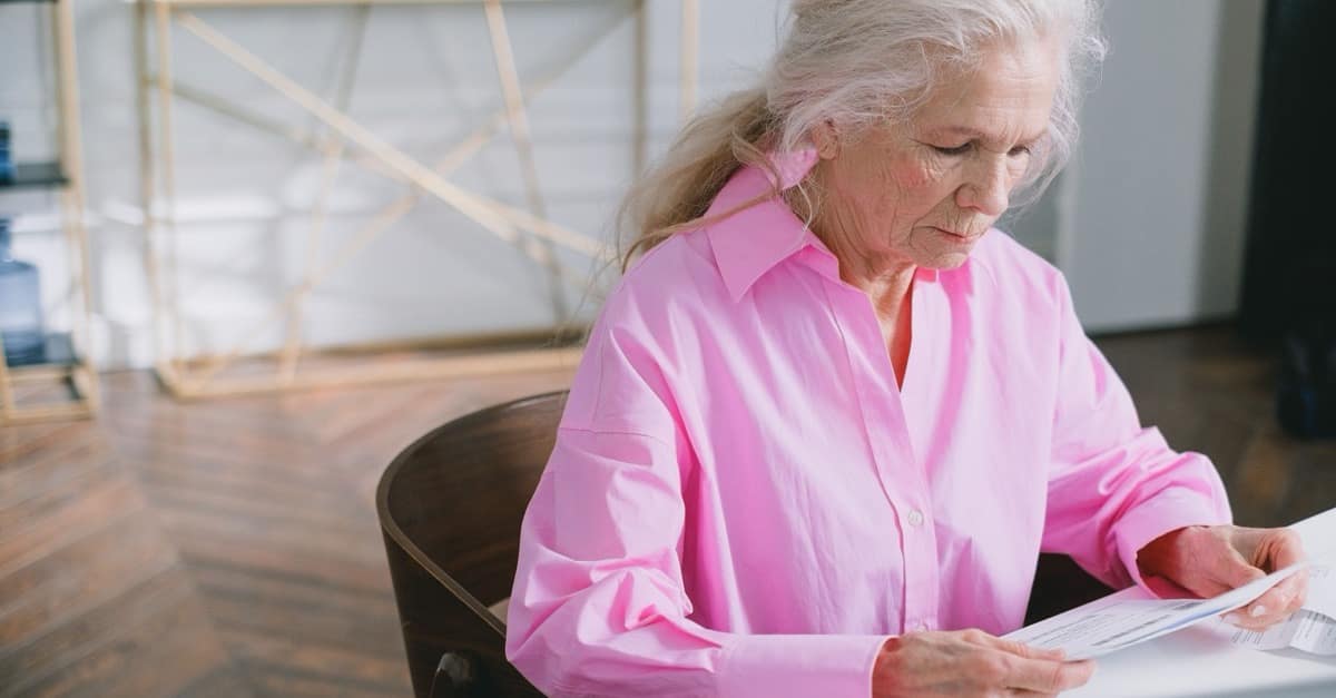 A grey-haired woman in her seventies sits at a white table in a bright room, reading a printed document.