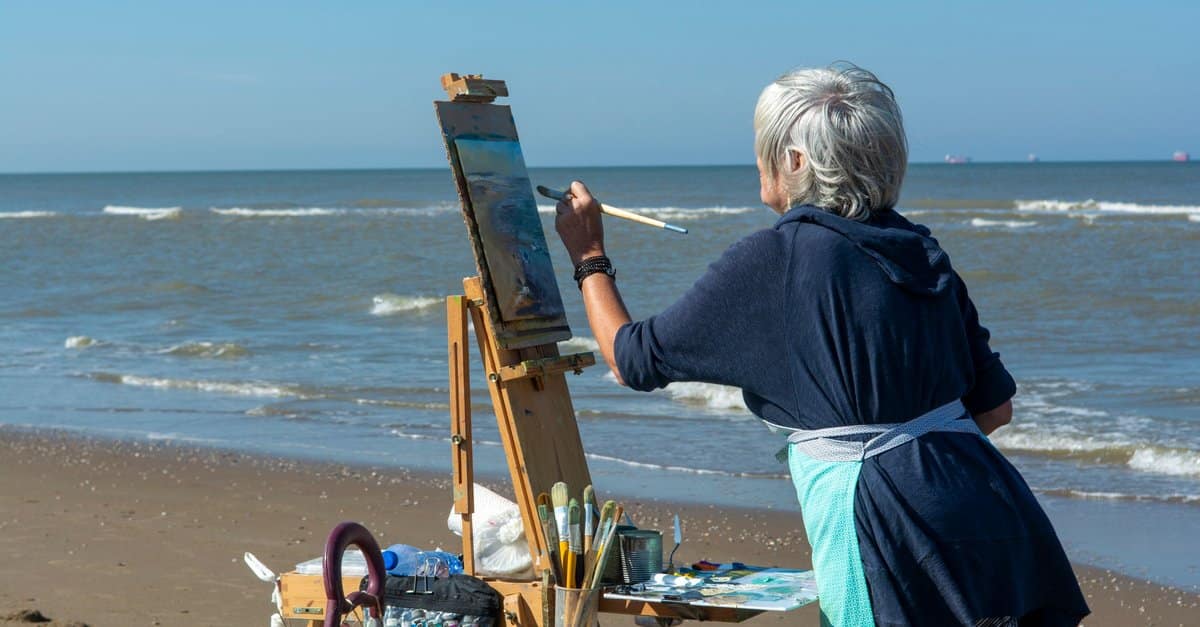 A grey-haired woman paints at an easel on the beach with the sea behind her.