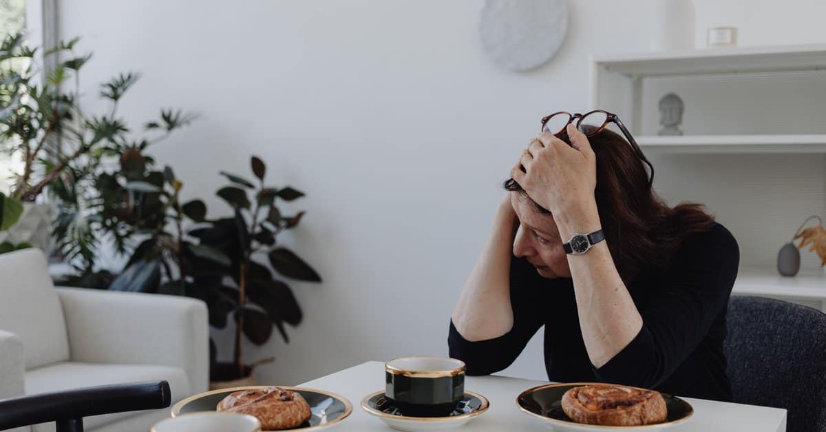 A woman sits at a table with her head in her hands, coffee and pastries untouched in front of her.
