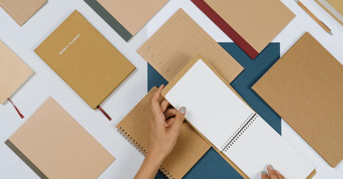 Overhead view of kraft-paper notebooks and a weekly planner laid out on a desk as a hand opens a spiral notepad.