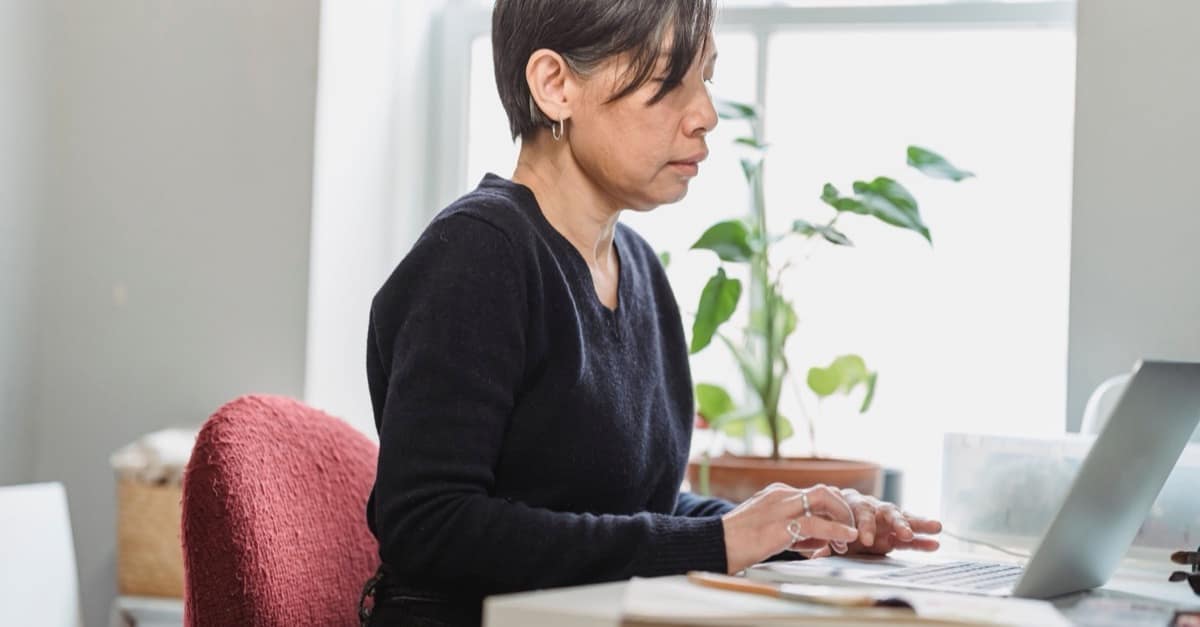 A woman with short dark hair types on a laptop at a desk by a bright window, calm and focused, with papers beside her.