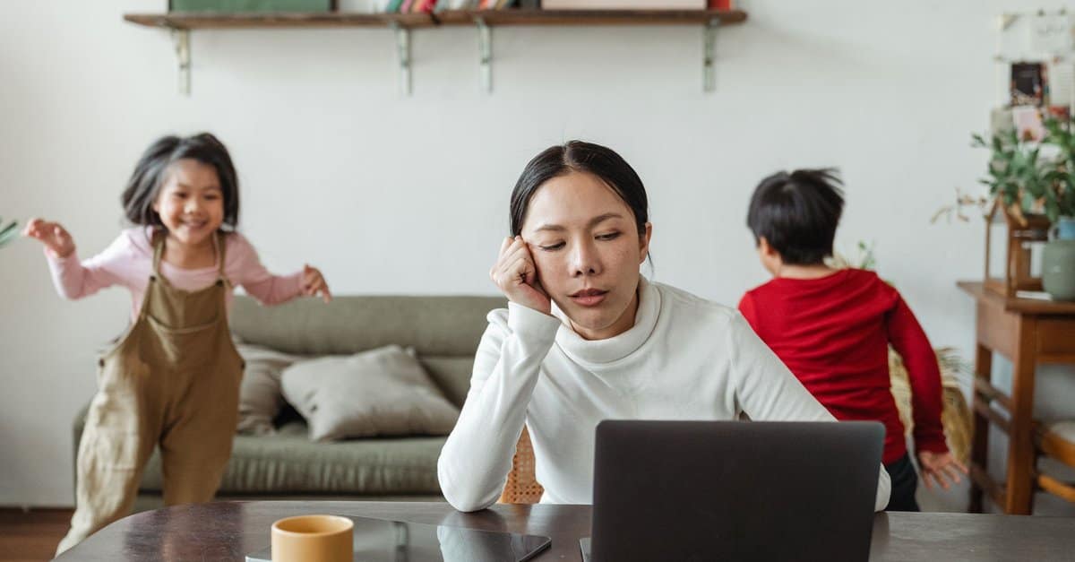 An exhausted mother rests her head on her hand at a laptop while her two children run around the living room.