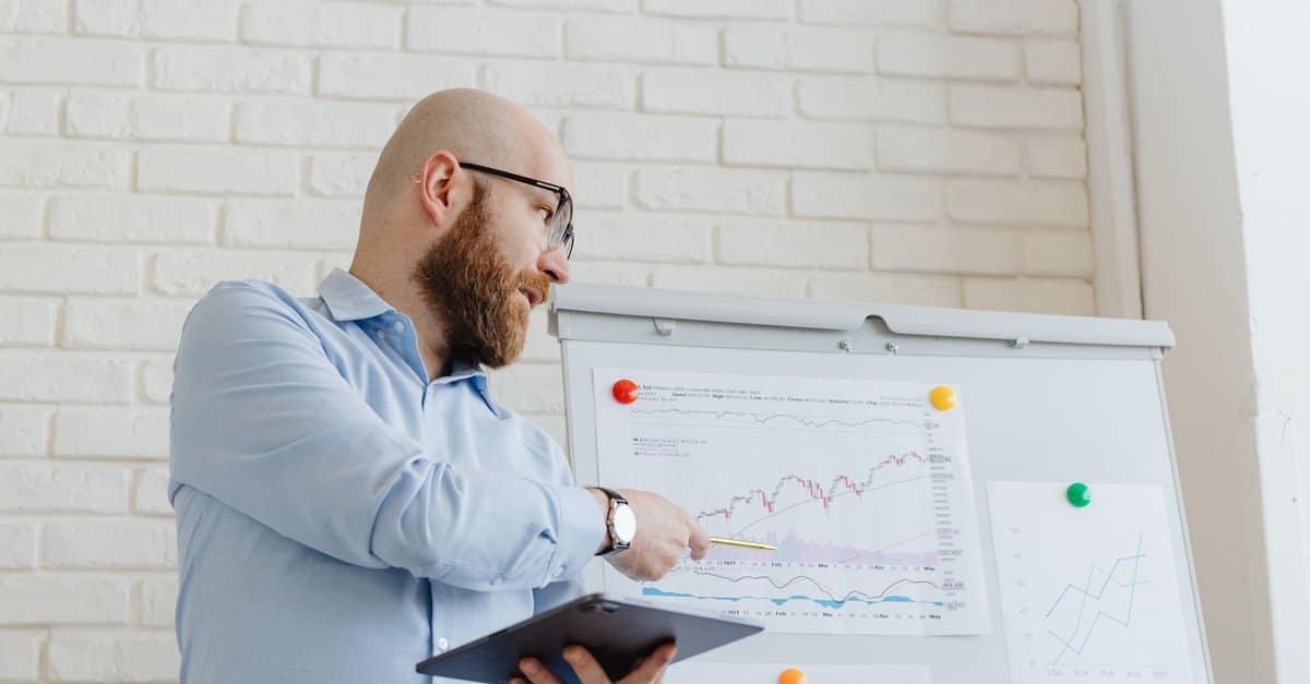 A man points to a performance chart on a whiteboard while holding a tablet during a work review.