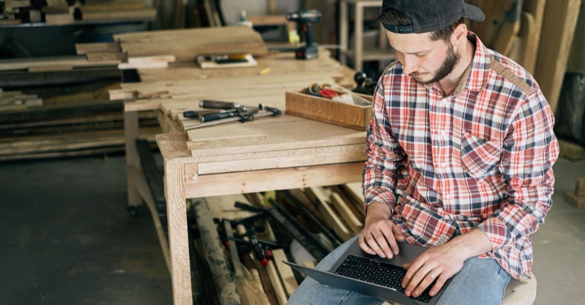 A carpenter uses a laptop at a workbench surrounded by tools and unfinished wood.