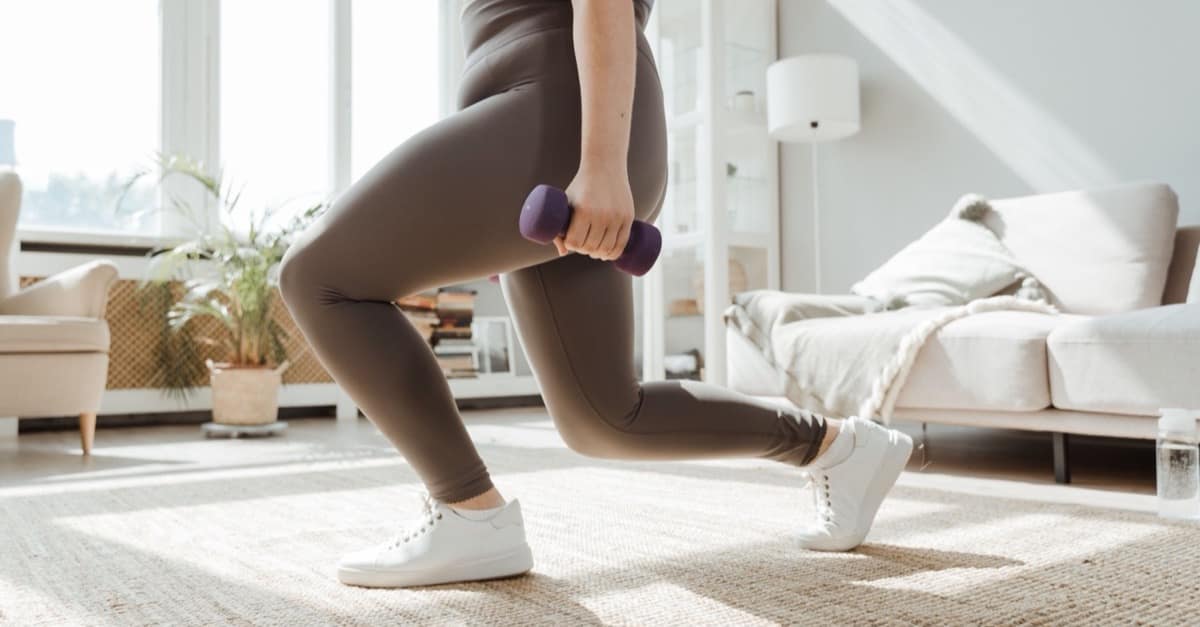 A woman holding dumbbells while lunging in a bright living room.