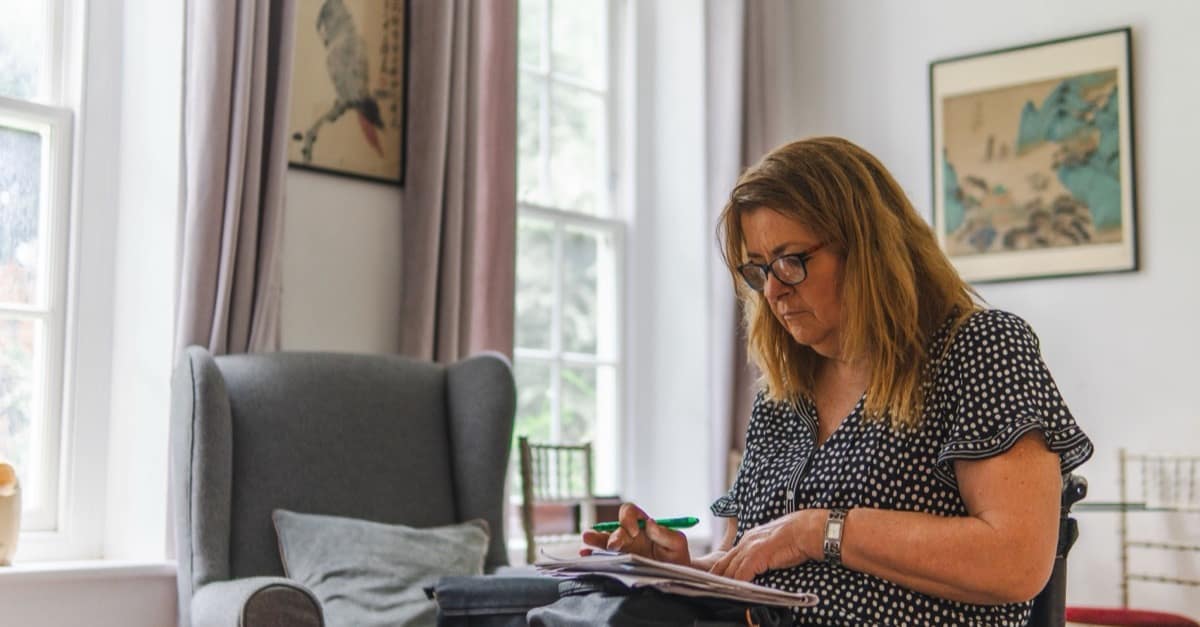 A woman in her fifties sits in a bright living room beside tall windows, writing notes with a green pen on papers in her lap.