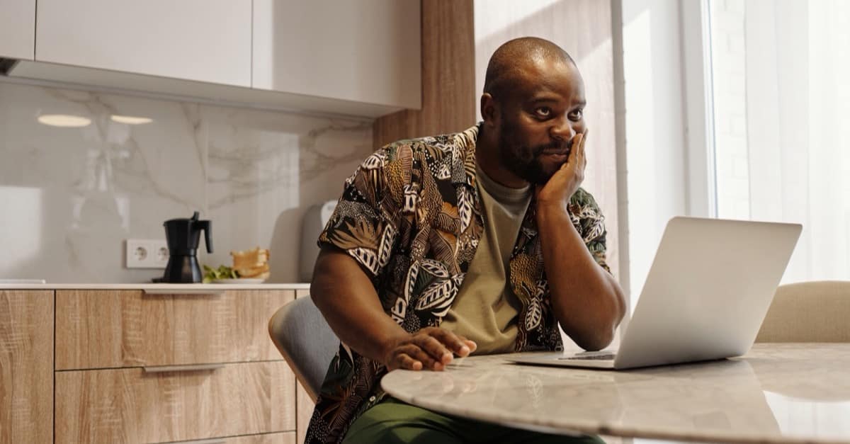 A man in a patterned shirt sits at a bright kitchen table with his chin in his hand, looking thoughtfully past his open laptop.