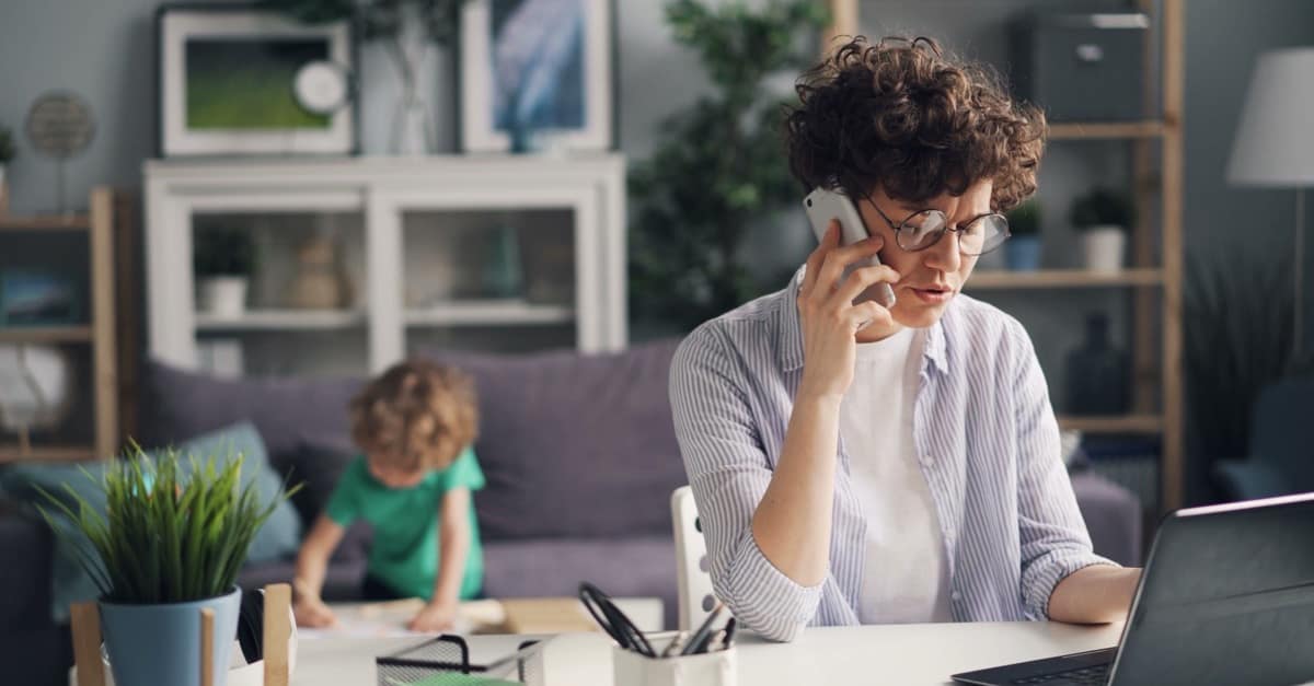 A mother talks on the phone at her desk beside an open laptop while her toddler plays on the sofa behind her in a bright living room.
