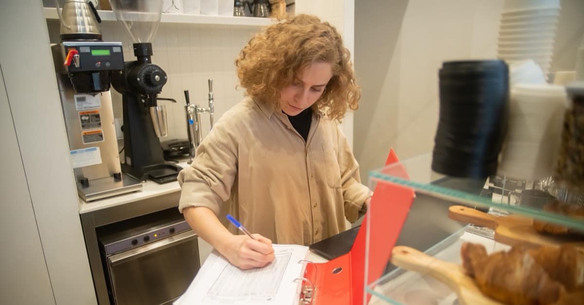 A small cafe owner in a beige shirt stands at her counter beside the espresso grinder, filling in a printed sales sheet from an open red binder, with croissants in the glass case in front of her.