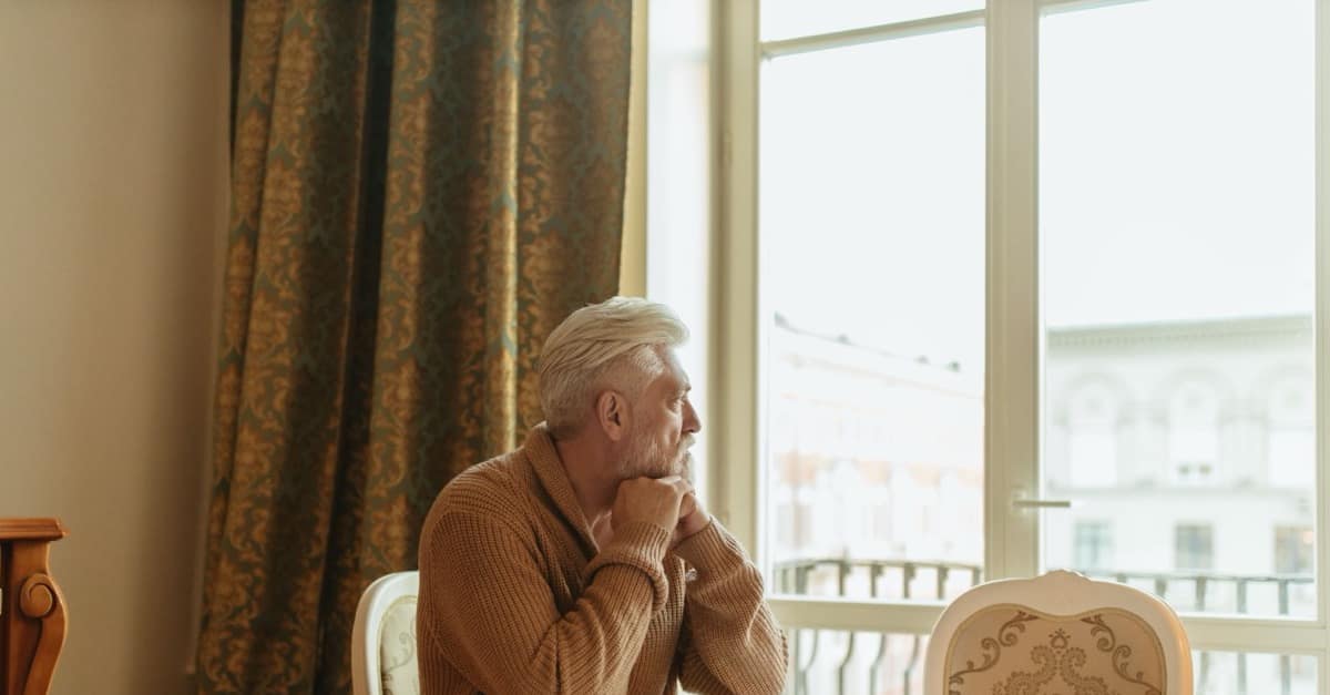 A gray-haired man in a brown sweater sits alone by a bright window, chin resting on his folded hands, looking outside.