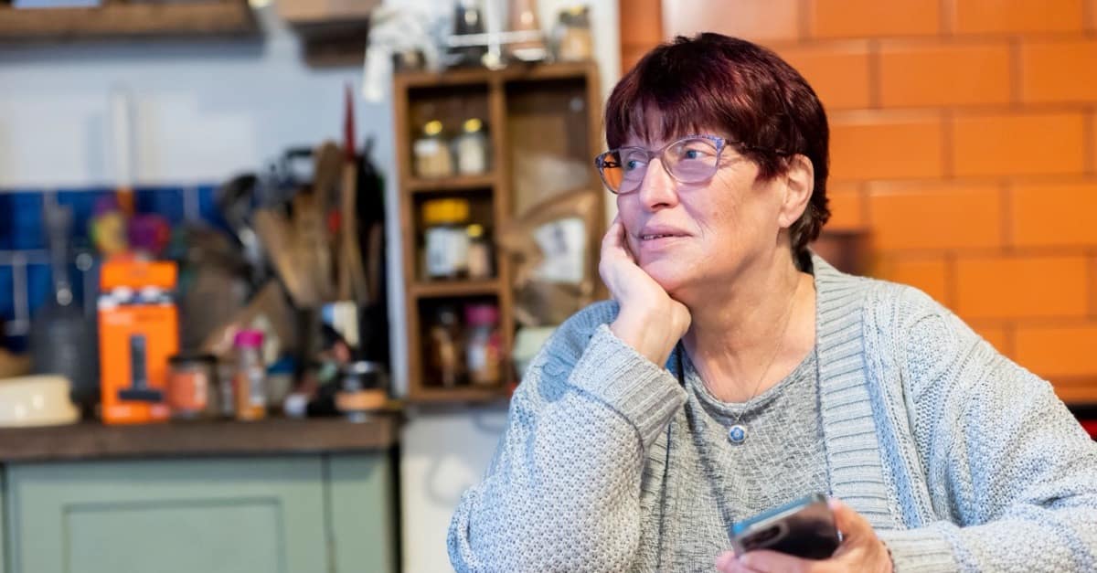 An older woman sits at her kitchen table with her phone in her hand and her chin resting on her palm, looking away toward the window.