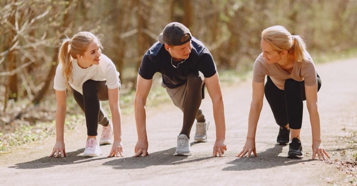 Three adults in running clothes crouch side by side at the start of a sunny park path, smiling at each other before a group run.