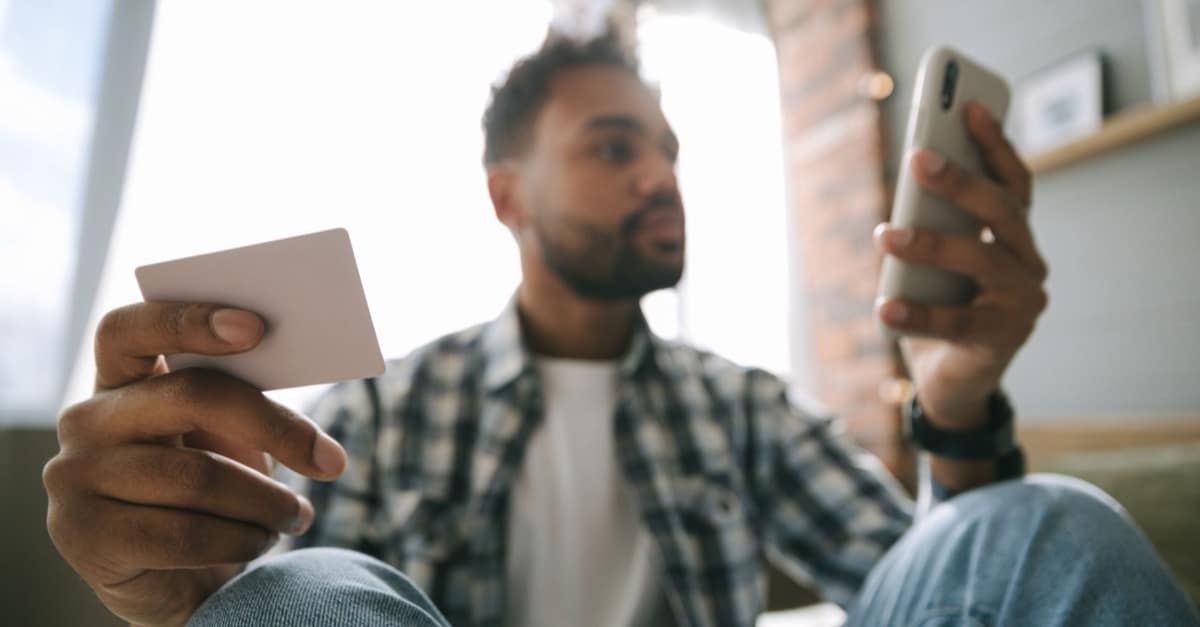 A man in a bright room holds a payment card in one hand and a smartphone in the other, about to buy something online.
