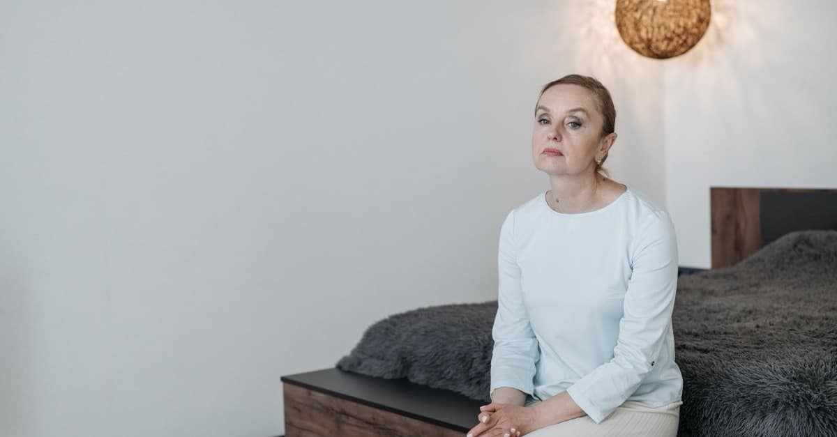 A woman in her fifties sits awake on the edge of her bed in a bright bedroom, hands folded, a pendant lamp glowing behind her.