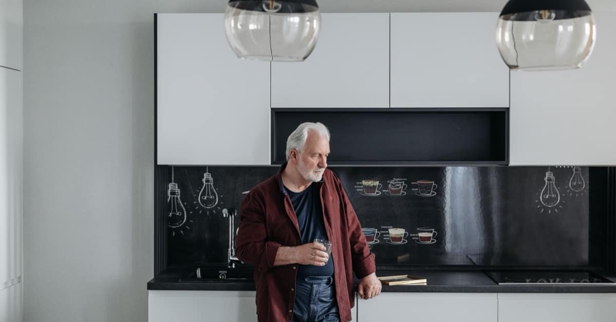 A gray-haired man in his sixties stands alone at his kitchen counter on a weekday morning, holding a glass and looking down.
