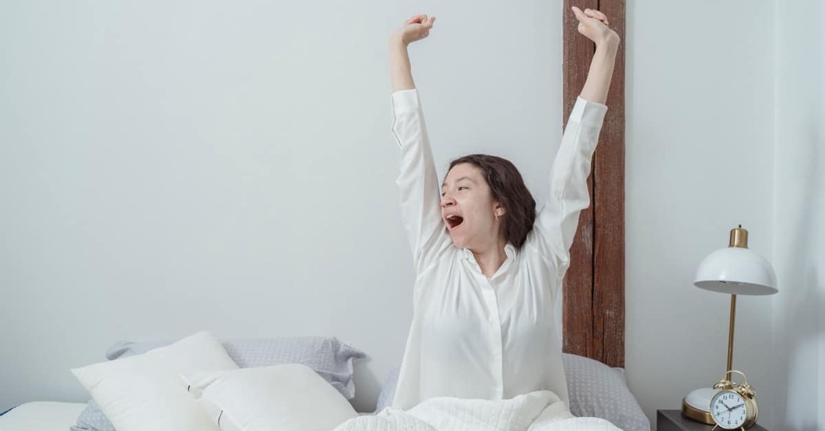 A woman in a white shirt sits up in bed in a bright bedroom, mid-yawn with both arms stretched overhead, an alarm clock on the nightstand beside her.