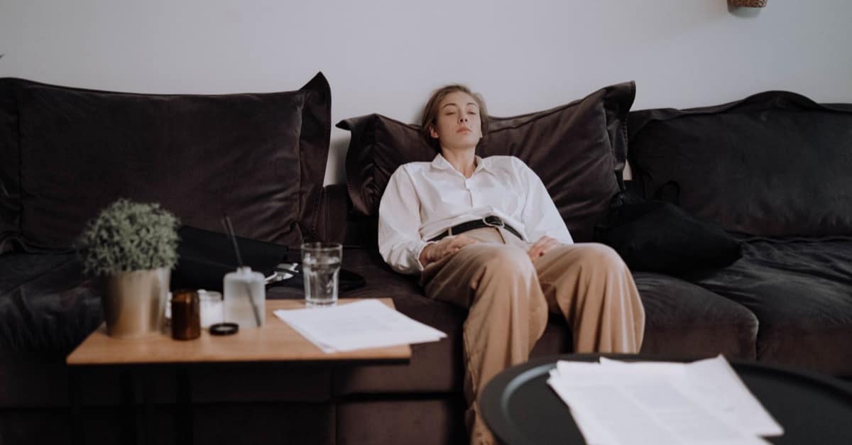 A woman in a white work shirt and trousers slumped back on a dark sofa with her eyes closed, papers and a glass of water on the table beside her.