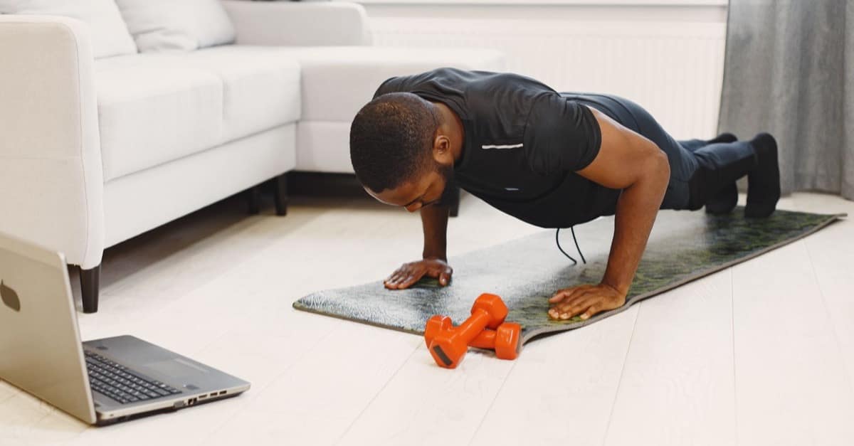 A man in black workout clothes does a push-up on a mat in a bright room, a laptop open on the floor beside him.
