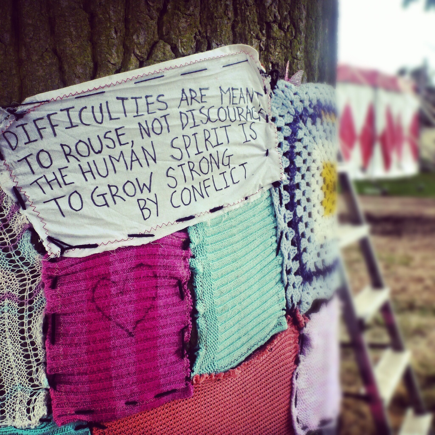 Tree trunk covered with knitting and material with the quote 'Difficulties are meant to rouse, not discourage. The human spirit is to grow strong by conflict.' Installation by Lunar Rising at Shambala Festival