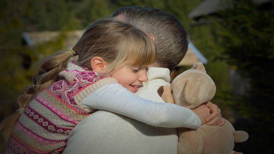 Image of a little gurl holding a teddybear and hugging her father