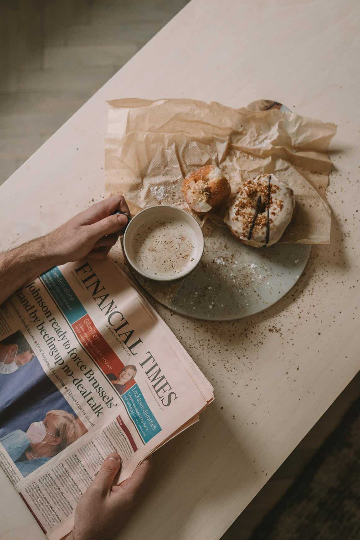 man reading newspaper on the table with a cup of coffee on hand and donuts on the plate